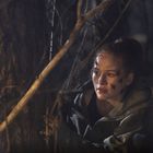 Eine Frau mit Schrammen im Gesicht kauert im Wald hinter einem Baum und schaut konzentriert nach vorne.