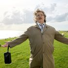 Ein Mann in einem beigen Mantel steht mit ausgestreckten Armen auf einem Deich, hinter ihm Schafe und Wasser.