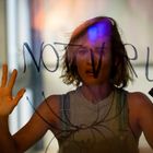 Frau hinter Glas, Text "NOT YET", blickt ernst, Hand erhoben, Hintergrund verschwommen.