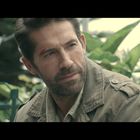 Ein Mann mit Bart und leichter Verletzung am Kopf trägt eine khakifarbene Jacke; im Hintergrund grüne Blätter.