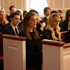 Eine Gruppe von Menschen in formeller Kleidung sitzt auf hölzernen Bänken in einem Raum mit farbigen Fenstern.