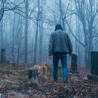 Ein nebliger Wald mit kahlen Bäumen, Gräbern, einer Person mit Jacke und einem Hund auf einem Friedhof.