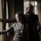 Zwei Personen in historischer Kleidung, neben einem Fenster stehend. Der Mann hält einen Hut und zeigt nach draußen.
