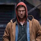 Ein Mann mit entschlossenem Ausdruck trägt eine orangene Kapuzenjacke. Der Hintergrund zeigt unscharfe Treppenstufen.