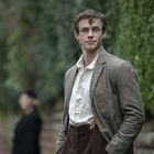 Ein Mann in brauner Jacke und weißem Hemd steht draußen, im Hintergrund ist eine unscharfe Person zu sehen.
