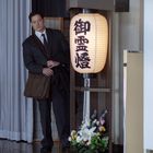 Eine Person im Anzug steht nachdenklich mit einer Tasche neben einem Aufsteller mit japanischen Schriftzeichen und Blumen.