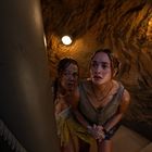 Zwei Frauen stehen erschrocken in einer spiralförmigen Treppe aus Stein, beleuchtet von Wandlampen.