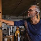 Ein Mann mit Brille malt konzentriert an einem Gemälde, umgeben von Ateliergegenständen und Skulpturen.
