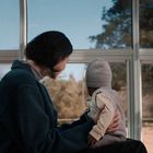 Kein Text im Bild. Eine Frau hält ein Baby auf dem Schoß und schaut aus einem großen Fenster in einen Garten.