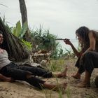 Ein Mann sitzt im Sand, während eine Frau hockt und ein Messer hält. Beide sind in einer dschungelartigen Umgebung.