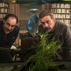 Zwei Männer betrachten ein Aquarium mit bunten Fischen; im Hintergrund sind viele Bücherregale zu sehen.