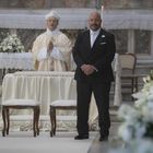 Zwei Männer vorne in einem prunkvollen Raum; einer trägt priesterliche Gewänder, der andere einen schwarzen Anzug.