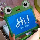 Ein Bildschirm zeigt "Hi! Let's play!" in einem grünen Froschrahmen. Spielzeugfiguren liegen auf einem Holzboden daneben.