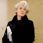 Eine elegante Frau mit weißem Haar trägt einen schwarzen Pelzmantel. Sie hält eine Mappe mit bunten Tabs und eine Brille.