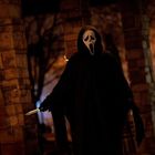 Eine Gestalt in dunklem Umhang mit gespenstischer Maske, hält ein Messer, umgeben von Säulen bei Nacht.