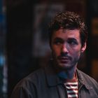 Ein Mann mit dunklen lockigen Haaren und Bart schaut nach rechts. Er trägt eine gestreifte Kleidung und steht vor unscharfem Hintergrund.