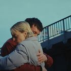 Zwei Menschen in dicken Jacken umarmen sich innig, im Hintergrund ist ein Geländer zu sehen.