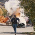 Es sind keine Texte vorhanden. Ein Mann rennt weg von einer Explosion, hinter ihm ein Motorrad und Rauch, umgeben von Bäumen.