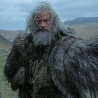 Es gibt keinen sichtbaren Text. Ein älterer Mann mit langem grauen Haar und Bart trägt einen dicken, zotteligen Mantel. Die Landschaft ist karg und hügelig.