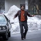 Ein Mann in brauner Lederjacke mit Polizeimarke steht vor einem alten Auto. Hintergrund: Schnee und Gebäude.