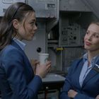 Zwei Personen in blauen Uniformen stehen in einer Flugzeugküche. Eine hält einen Becher, die andere hat die Arme verschränkt.