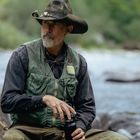 Ein Mann mit Hut sitzt am Fluss, trägt eine Anglerweste und hält eine Flasche in der Hand. Ruhige Naturkulisse im Hintergrund.