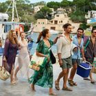 Text: "CATAMARAN". Eine Gruppe von Menschen geht lächelnd mit Strandtaschen und Kühlbox an einem Hafen entlang.
