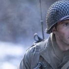 Ein Soldat mit Helm schaut ernst, trägt eine olivgrüne Uniform und hat einen Funkgerät-Antennendraht am Helm.