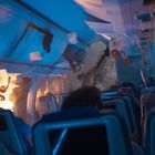 Keine sichtbaren Texte. Eine verwüstete Flugzeugkabine, herunterhängende Sauerstoffmasken, Chaos und Feuer.