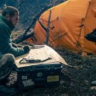 Ein Mann in Outdoor-Kleidung sitzt vor einem großen deutschen Schäferhund und einem Campingzelt, sie scheinen zu kommunizieren.
