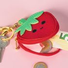 Eine kleine rote Geldbörse in Form einer Erdbeere mit Gesicht, gefüllt mit einem 5-Euro-Schein und Münzen, daneben ein Schlüssel.