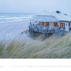 1 · 2027. Ein Strandhaus auf stilisierten Stelzen am Meer, umgeben von Dünen und Gräsern, bei bewölktem Himmel.