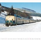 221 141-5. Ein Zug fährt durch eine verschneite Landschaft. Im Hintergrund Wald und Hügel, blauer Himmel.