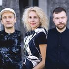 Drei Personen in modischer Kleidung vor unscharfem Hintergrund: links Mann mit weißer Mütze, Mitte Frau mit lockigem Haar.