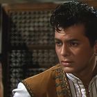 Eine Person mit ernstem Gesichtsausdruck trägt traditionelle Kleidung und Ohrringe; im Hintergrund ein dekorativer Paravent.