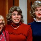 Text: "KEEP IT UP". Drei Personen mit Perücken, femininer Kleidung und Make-up lächeln in einer Holzvertäfelung.