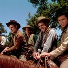 Fünf Männer auf Pferden, in Westernkleidung, tragen Hüte, lächeln. Hintergrund: Bäume und blauer Himmel.