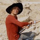 Person mit großem Hut und rotem Pulli hockt in sandiger Landschaft, hält ein Gewehr in der Hand und blickt ernst.