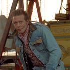 Ein Mann in Jeansjacke und gemustertem Halstuch lehnt sich vor einem gelben Karussell mit Adler-Dekor nach vorne.