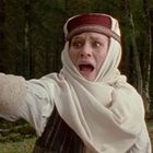 Eine Person im weißen Gewand und Hut zeigt schockiert mit dem Finger; sie steht in einem Wald.