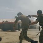 Drei uniformierte Personen in militärischen Westen laufen durch eine staubige Umgebung, eine davon führt eine Geisel.