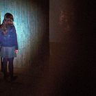 Ein Mädchen mit langem Haar steht vor einer großen, dunklen Tür in einem schwach beleuchteten Raum.