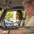 Zwei Männer sitzen in einem Fahrzeug, der eine am Steuer. Draußen sind Autos und Bäume zu sehen.