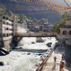 Viele bunte Gebetsfahnen spannen sich über einen Fluss. Gebäude umgeben das Wasser; Menschen baden im Dampf eines Beckens.