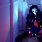 Eine Person in Schuluniform sitzt mit einer Maske in einem Flur mit violettem Licht.