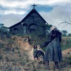 Person in langem Mantel und Kapuze steht mit Hund vor einer alten Holzkirche auf einem hügeligen, bewachsenen Gelände.