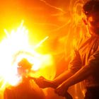 Keine erkennbaren Texte. Ein Mann hält einen Flammenwerfer, der helle Funken versprüht. Alles in warmem, orangenem Licht.