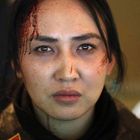 Eine Frau mit dunklen Haaren und ernstem Gesichtsausdruck, blutverschmiert auf Stirn und Nase. Hintergrund unscharf.