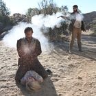 Zwei Männer in einer Wüstenlandschaft, einer kniet mit gefesselten Händen, der andere steht mit einer Pistole in der Hand.