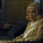 Keine sichtbaren Texte. Eine ältere Frau mit blonden Haaren sitzt auf einem Sofa in einem schwach beleuchteten Raum.